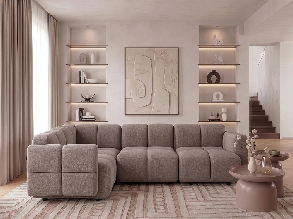 A modern sectional sofa in a warm taupe-gray fabric, styled in a contemporary living room with built-in shelves, a patterned rug, and minimalist tables.