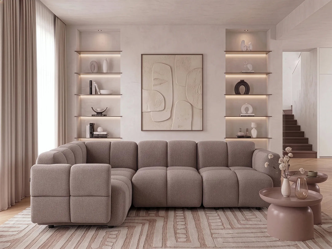 A modern sectional sofa in a warm taupe-gray fabric, styled in a contemporary living room with built-in shelves, a patterned rug, and minimalist tables.