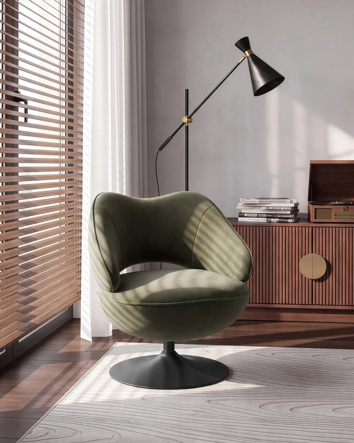 An olive green upholstered swivel armchair with a black pedestal base, styled in a modern room next to a window with wooden blinds, a black floor lamp, and a wooden cabinet.