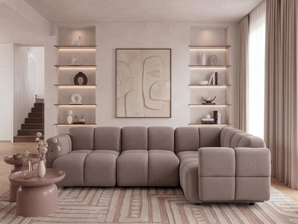 A modern sectional sofa in a warm taupe-gray fabric, styled in a contemporary living room with built-in shelves, a patterned rug, and neutral-toned curtains.