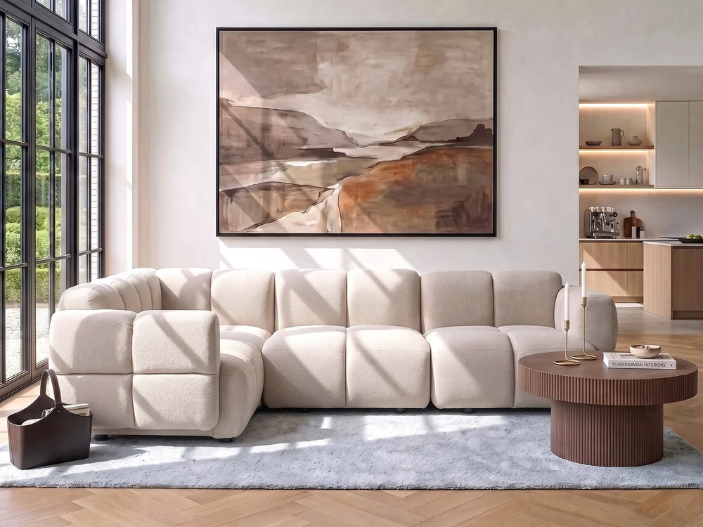 A modern sectional sofa in a warm off-white or cream upholstery, styled in a bright, contemporary living room with a large abstract landscape painting on the wall, a round wooden coffee table, and a light blue rug.
