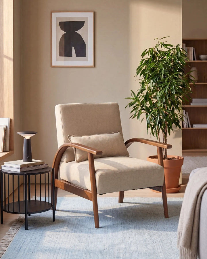 A beige upholstered armchair with curved wooden arms and legs, styled in a warm, modern living room setting with a blue rug, side table, potted plant, and framed wall art.