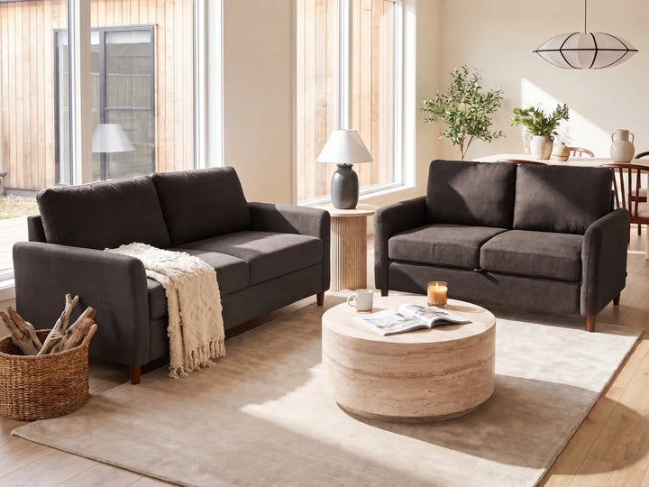 A dark gray fabric living room furniture set, including a loveseat and a matching armchair, arranged in a bright, modern living room with a round wooden coffee table and a beige rug.