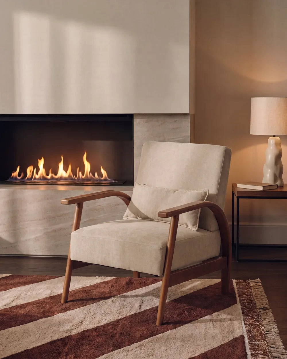 A mid-century modern upholstered armchair with a cream fabric and curved wooden arms, styled in a cozy living room next to a fireplace and a side table with a lamp.
