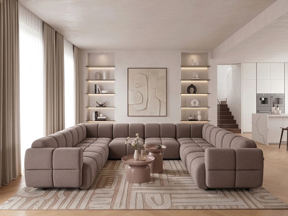 A large U-shaped modular sectional sofa in taupe fabric, styled in a modern, bright living room with a patterned rug, round coffee tables, and built-in shelving.