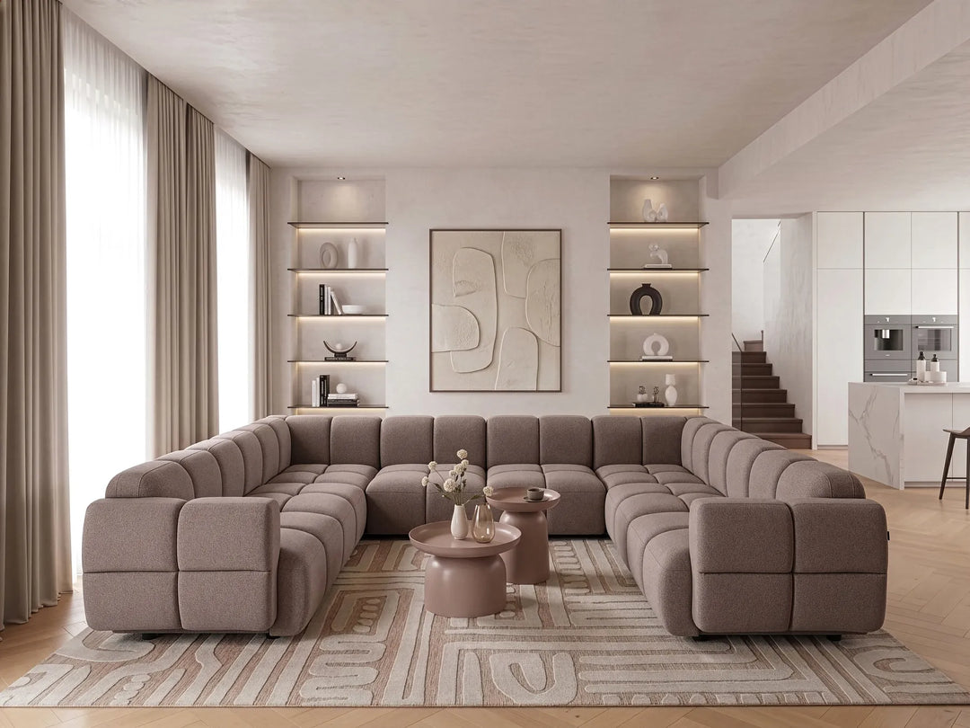 A large U-shaped modular sectional sofa in taupe fabric, styled in a modern, bright living room with a patterned rug, round coffee tables, and built-in shelving.