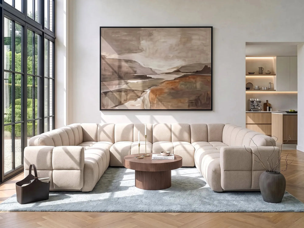 A large framed abstract landscape painting hangs on a white wall above a beige sectional sofa in a modern living room with wood floors, a light blue rug, and a round wooden coffee table.