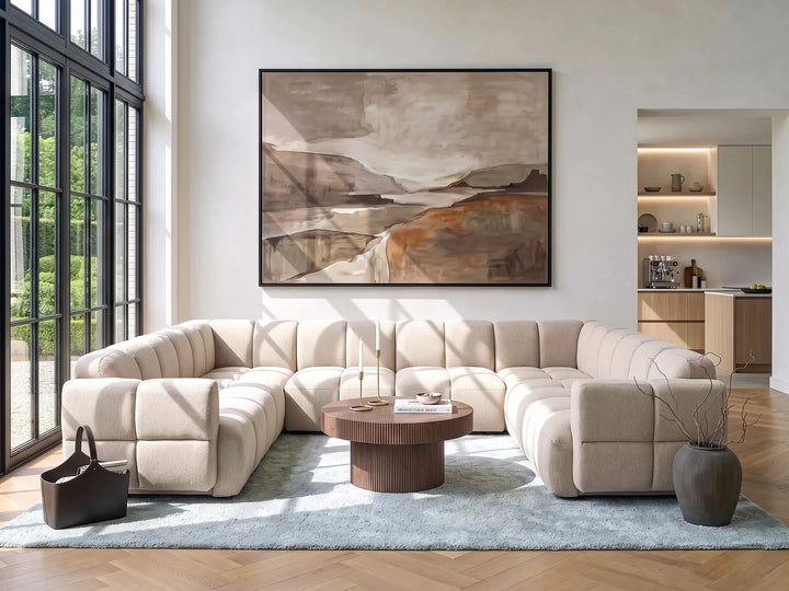 A large framed abstract landscape painting hangs on a white wall above a beige sectional sofa in a modern living room with wood floors, a light blue rug, and a round wooden coffee table.