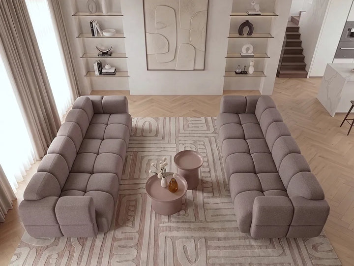 A modern living room setup featuring two tufted modular sofas in a muted taupe-gray color, arranged on a patterned rug with two matching side tables in the center.