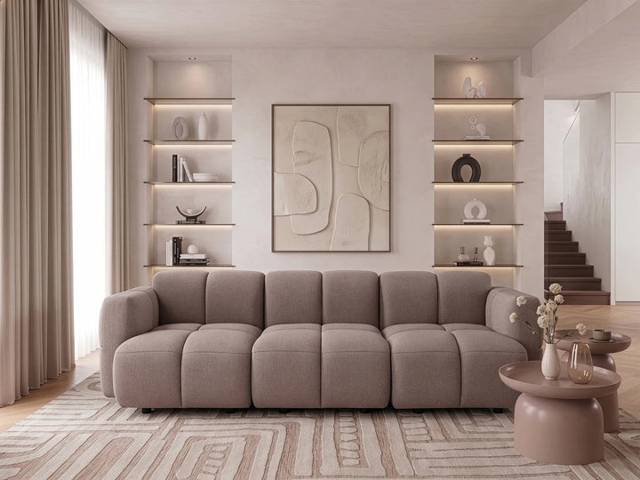 A modern modular sectional sofa in a warm taupe-gray fabric, styled in a minimalist, contemporary living room with built-in shelves, a patterned rug, and a matching side table.