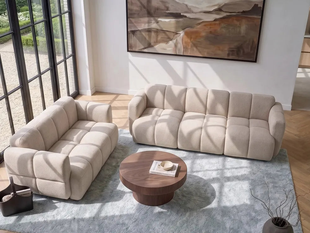 A modern living room setup featuring a beige tufted loveseat and a matching beige three-seater sofa arranged on a light blue-gray rug around a round wooden coffee table. Large black-framed windows on the left let in natural light, and an abstract painting hangs on the white wall above.