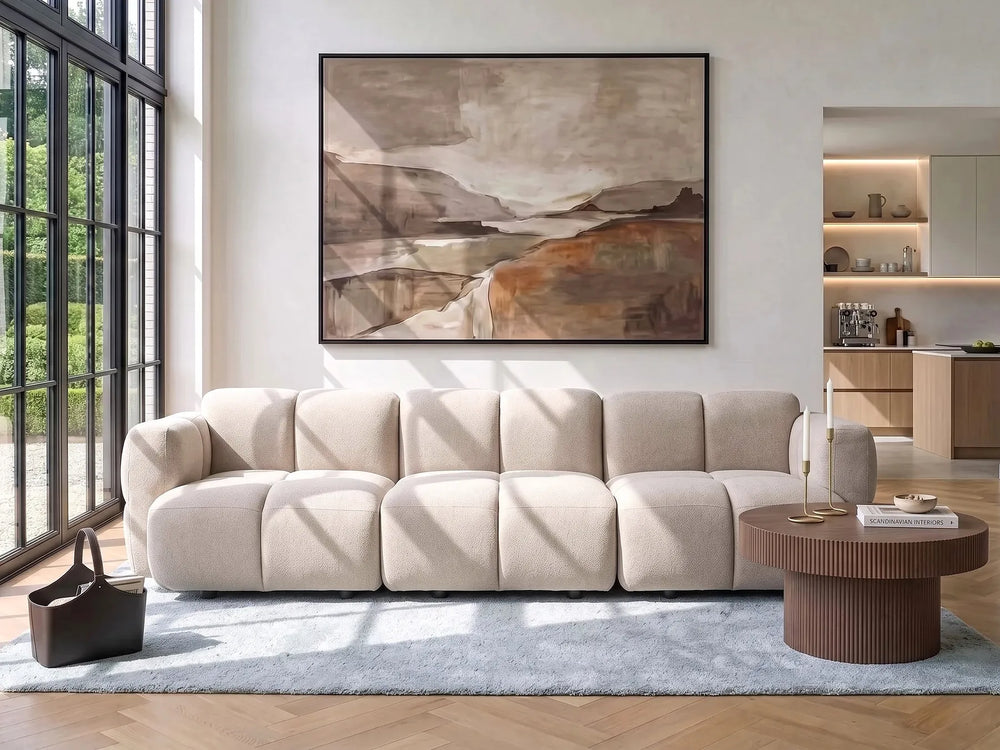 A modern beige modular sectional sofa in a bright, contemporary living room with a large abstract landscape painting on the wall, a round wooden coffee table, and a light blue rug.