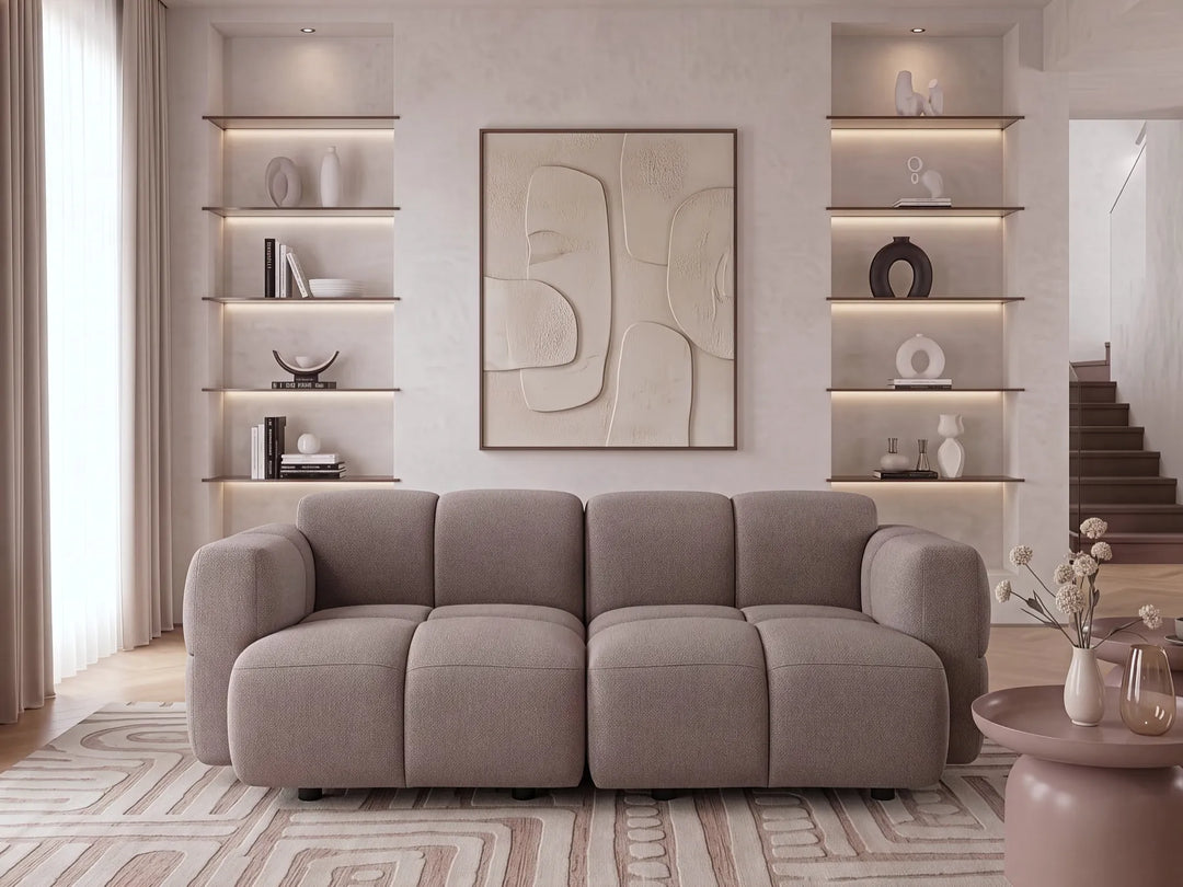 A modern three-seater sofa in a warm taupe-gray fabric, styled in a contemporary living room with built-in shelves, abstract wall art, and a patterned rug.