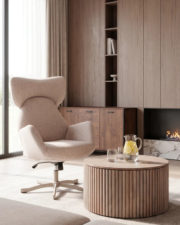 A modern beige upholstered accent chair with a high back and swivel base, placed in a styled living room next to a round ribbed wood coffee table and a fireplace.