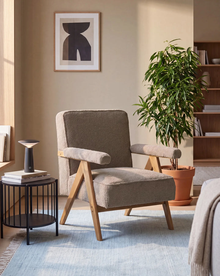 A mid-century modern armchair with beige textured upholstery and light wood legs, styled in a warm, neutral-toned living room next to a black wire side table and a potted plant.