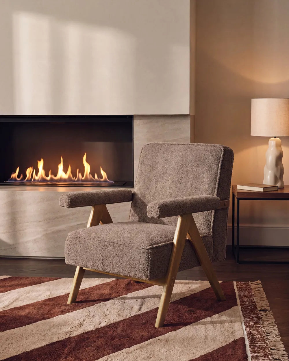A mid-century modern accent chair upholstered in textured taupe fabric with light wood legs, positioned in a cozy living room setting next to a fireplace and a side table with a lamp.