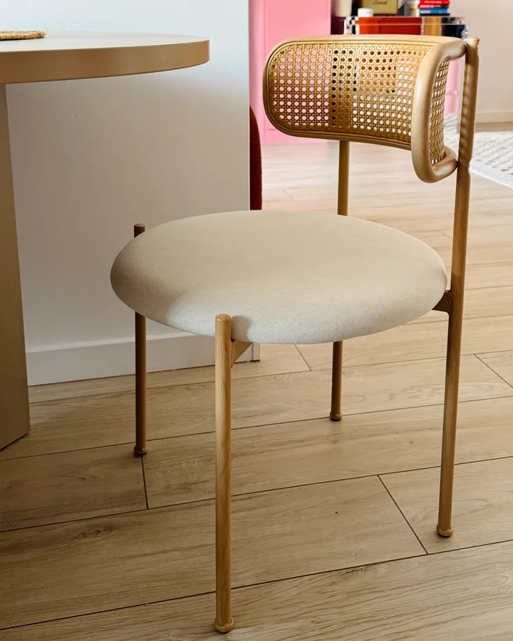 A modern dining chair with a light beige upholstered seat, a woven cane backrest, and a light wood frame, shown on a light wood floor next to a table.