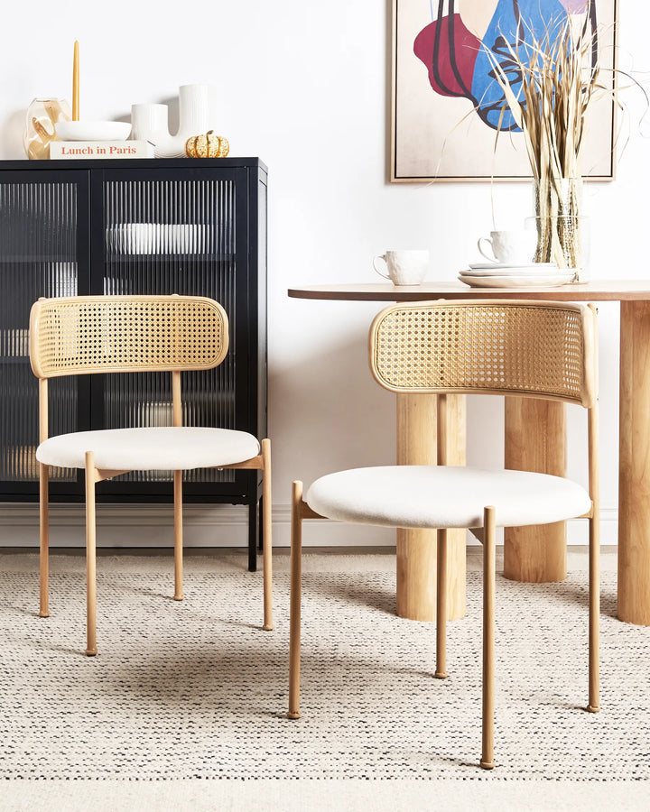 Two modern dining chairs with woven cane backs and cream upholstered seats, shown in a styled dining room setting next to a wooden table and cabinet.