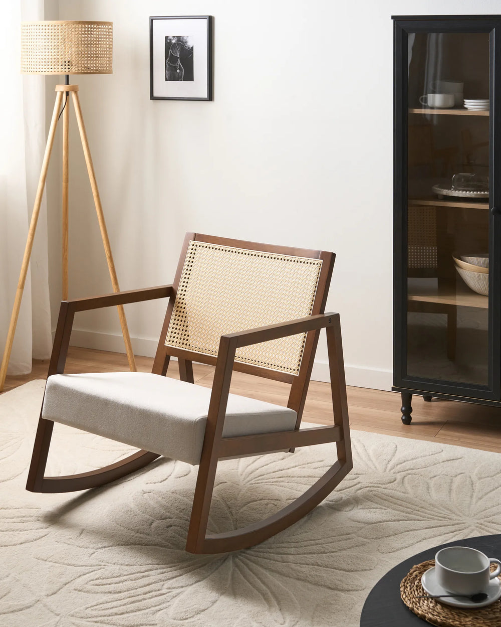A mid-century modern wooden rocking chair with a woven cane backrest and a light beige upholstered seat, styled in a bright, minimalist room next to a tripod floor lamp and a dark glass cabinet.