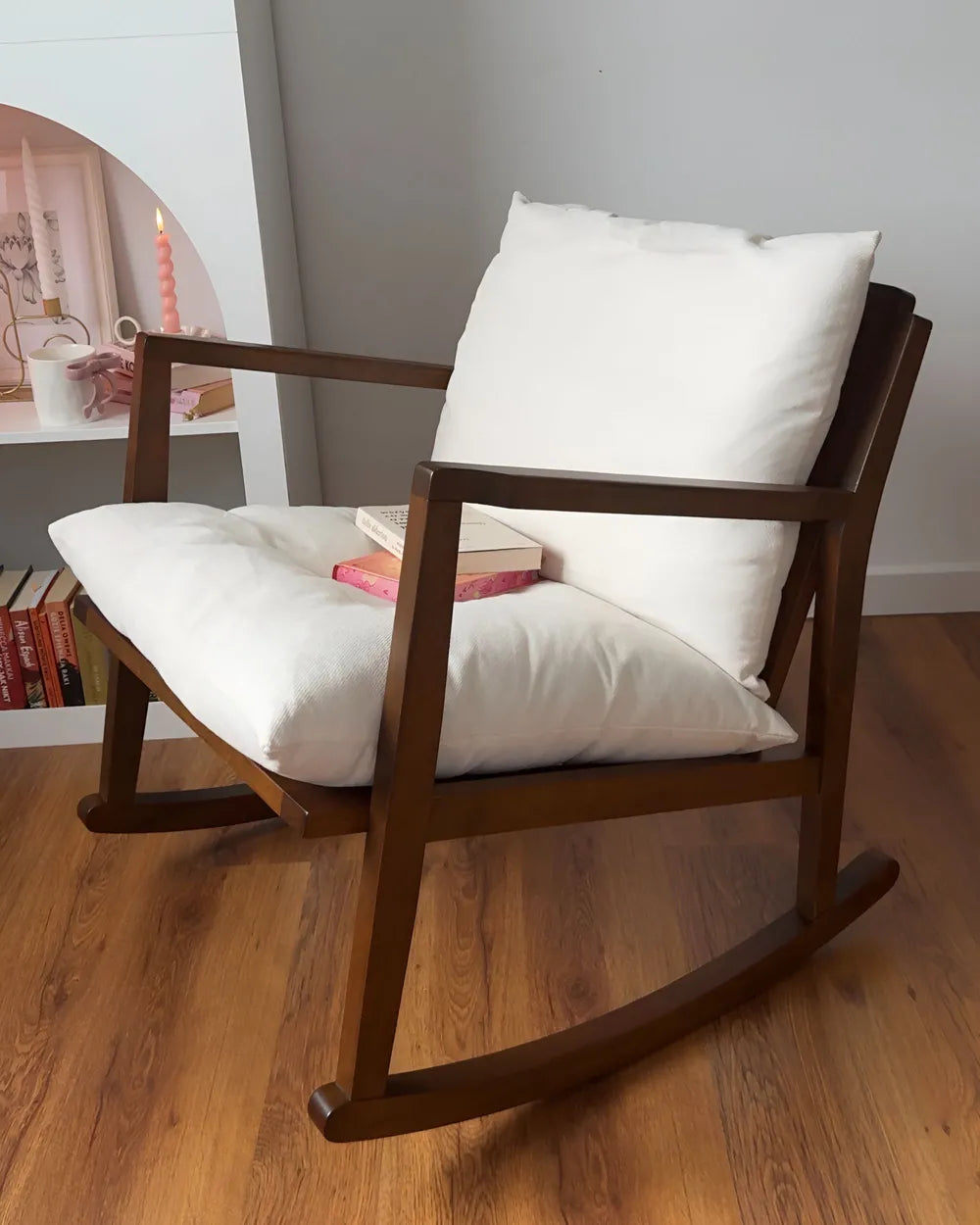 A mid-century modern wooden rocking chair with thick white cushions, styled on a warm wood floor next to a white shelf.