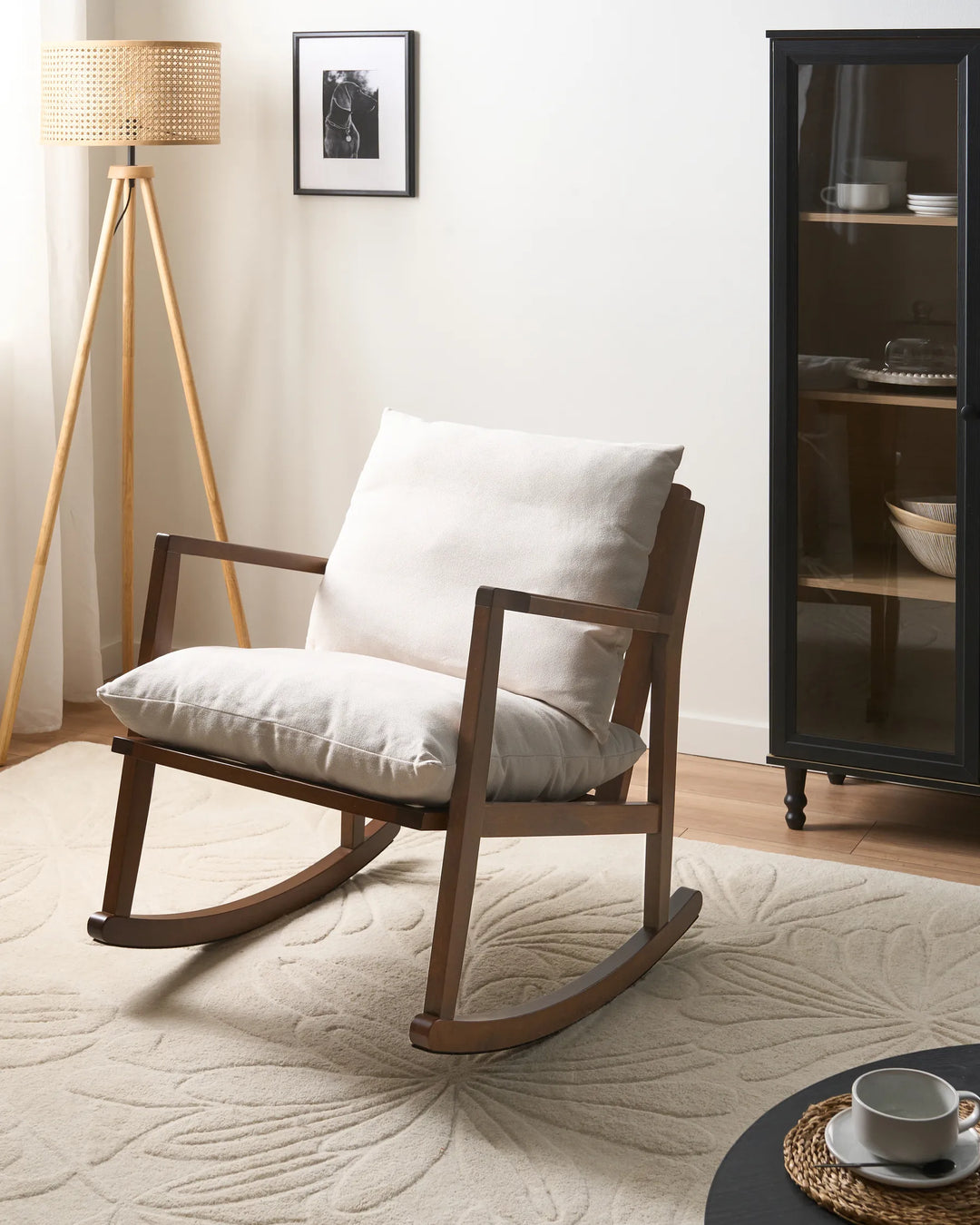 A mid-century modern wooden rocking chair with a white cushion, styled in a bright, minimalist room with a beige rug, a tripod floor lamp, and a black glass cabinet.