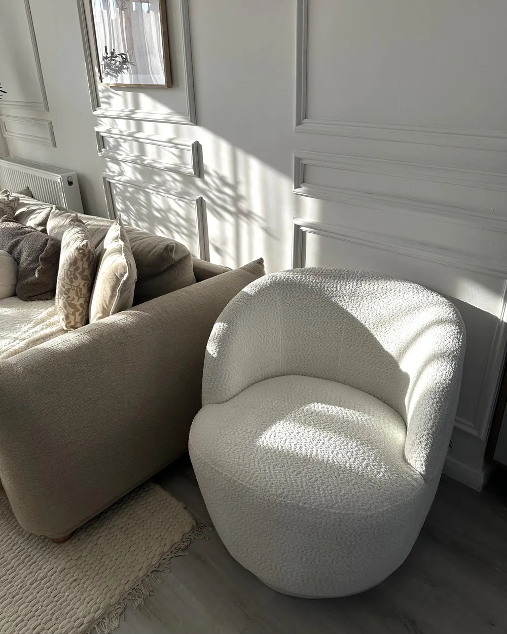 A modern, rounded boucle armchair in off-white textured fabric, positioned next to a beige sofa in a brightly lit living room with white paneled walls and natural sunlight.