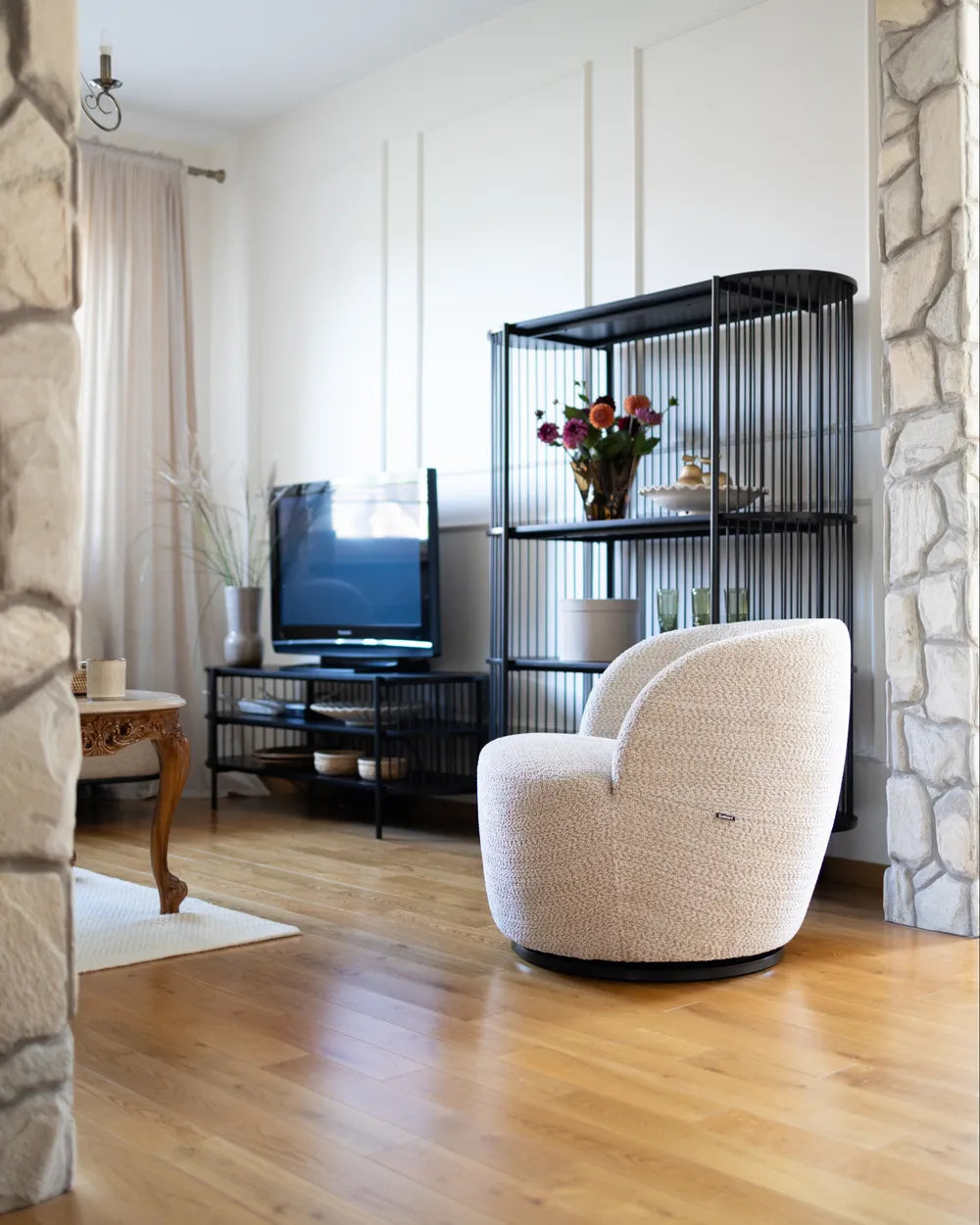 A modern cream bouclé swivel armchair on a black swivel base, set in a contemporary living room with light wood floors, a black wire shelving unit, and a television.