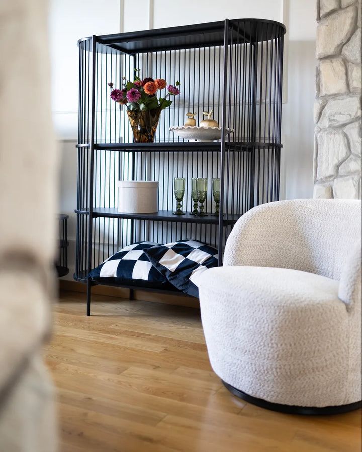 A black metal birdcage-style shelving unit with three tiers, styled in a living room setting with a white boucle armchair, wood floors, and a stone wall.