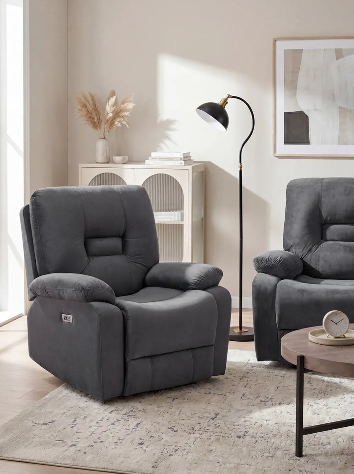 A dark gray upholstered recliner chair shown in a styled living room setting with a matching armchair, floor lamp, rug, and wall art.