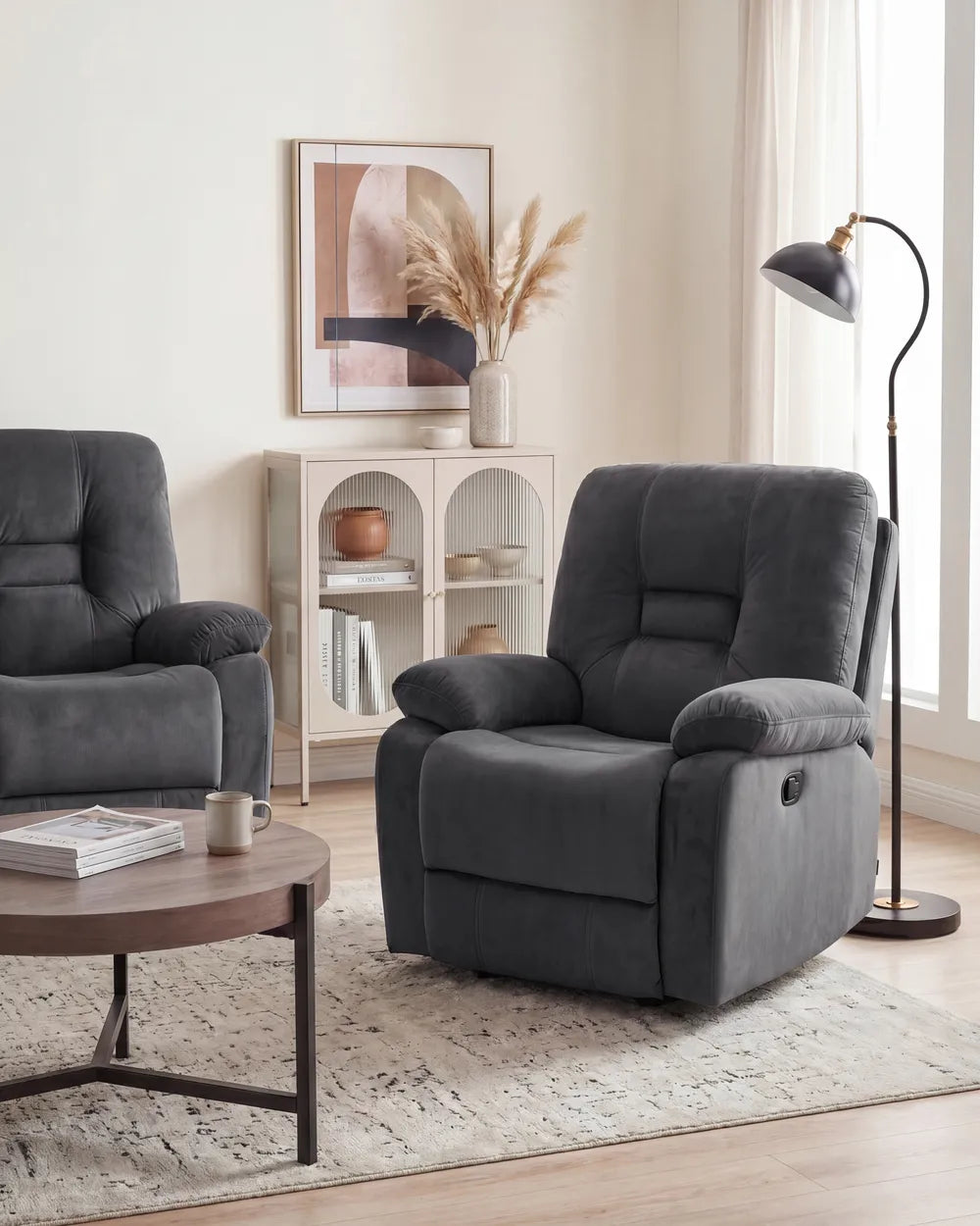 A dark gray upholstered recliner chair shown in a styled living room with a matching armchair partially visible, a round coffee table, a floor lamp, and a small cabinet in the background.