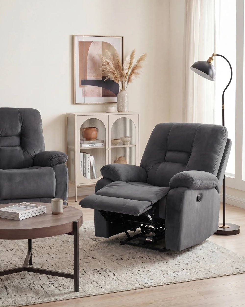 A dark gray recliner chair with its footrest extended, shown in a bright, modern living room setting next to a matching armchair, a coffee table, and a floor lamp.