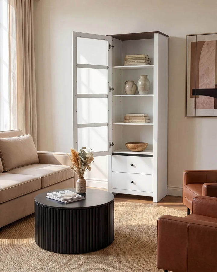 A tall white storage cabinet with glass-paneled doors and open shelves stands in a styled living room beside a beige sofa and round black coffee table.