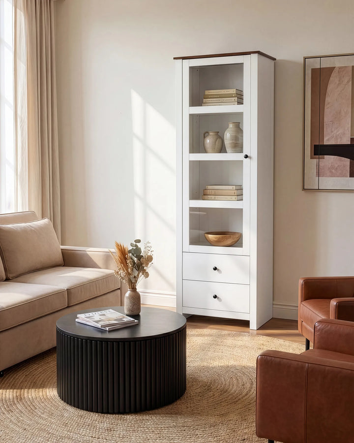 A tall white shelving cabinet with open upper shelves and two lower drawers, shown in a styled living room next to a beige sofa and round black coffee table.