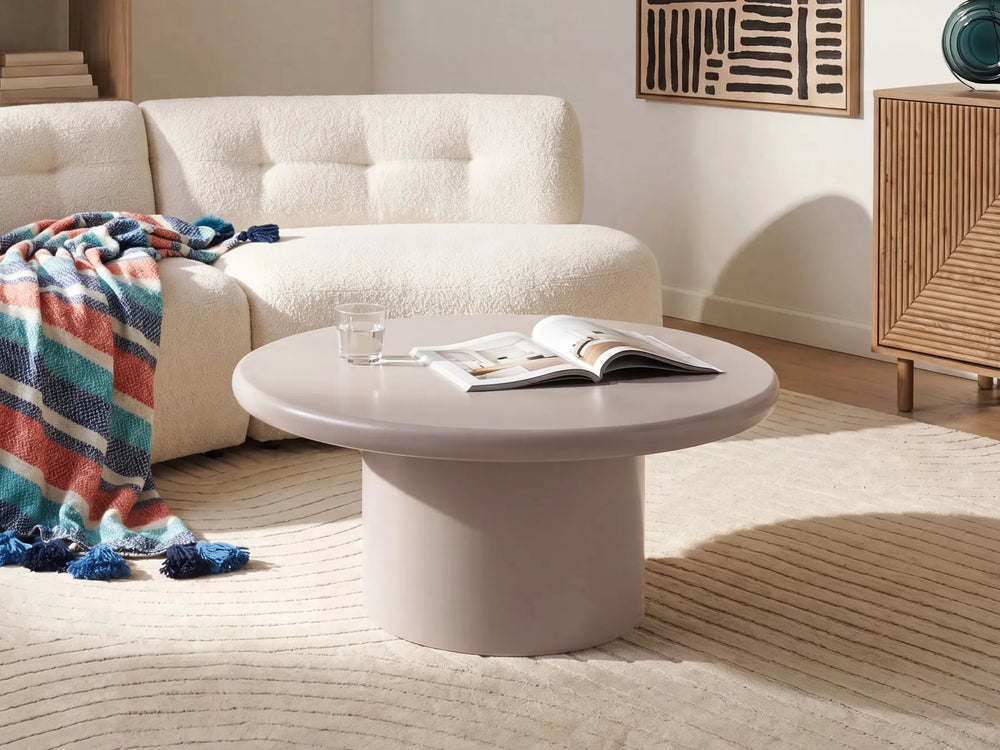 A round, low-profile coffee table in a light blush pink color, styled in a modern living room setting with a cream boucle sofa, a striped throw blanket, and a striped rug.