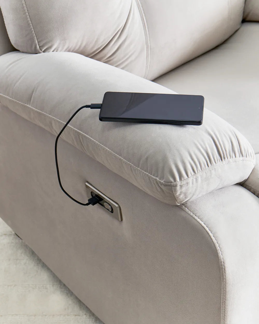 Close-up of a light beige upholstered armchair with a power outlet on the side, showing a black smartphone plugged in with a charging cable.