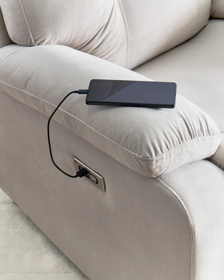 Close-up of a light beige upholstered armchair with a power outlet on the side, showing a black smartphone plugged in with a charging cable.