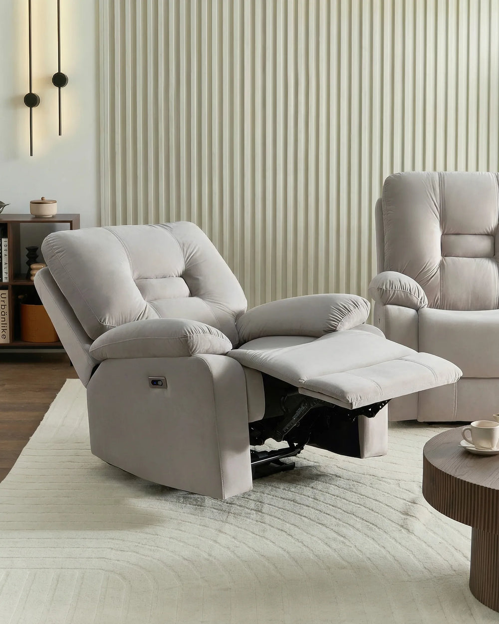A light gray upholstered recliner chair with the footrest extended, shown in a modern living room setting with a matching armchair, striped wall, and textured rug.