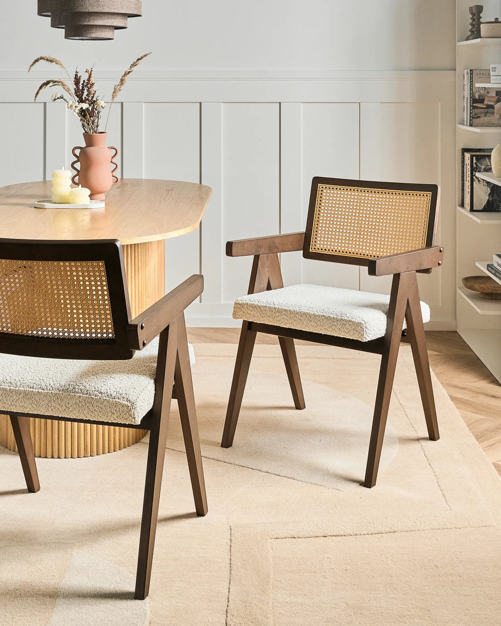 Two mid-century modern dining chairs with dark wood frames, woven cane backrests, and cream bouclé seat cushions, shown in a bright dining room setting next to a wooden table.