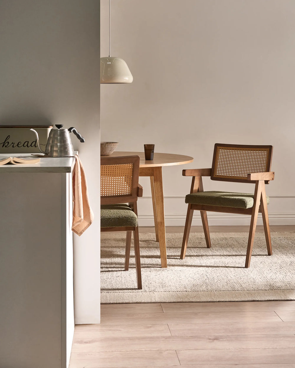 A mid-century modern wooden dining chair with a woven cane backrest and green cushioned seat, shown in a styled dining room setting next to a round wooden table and a light-colored rug.