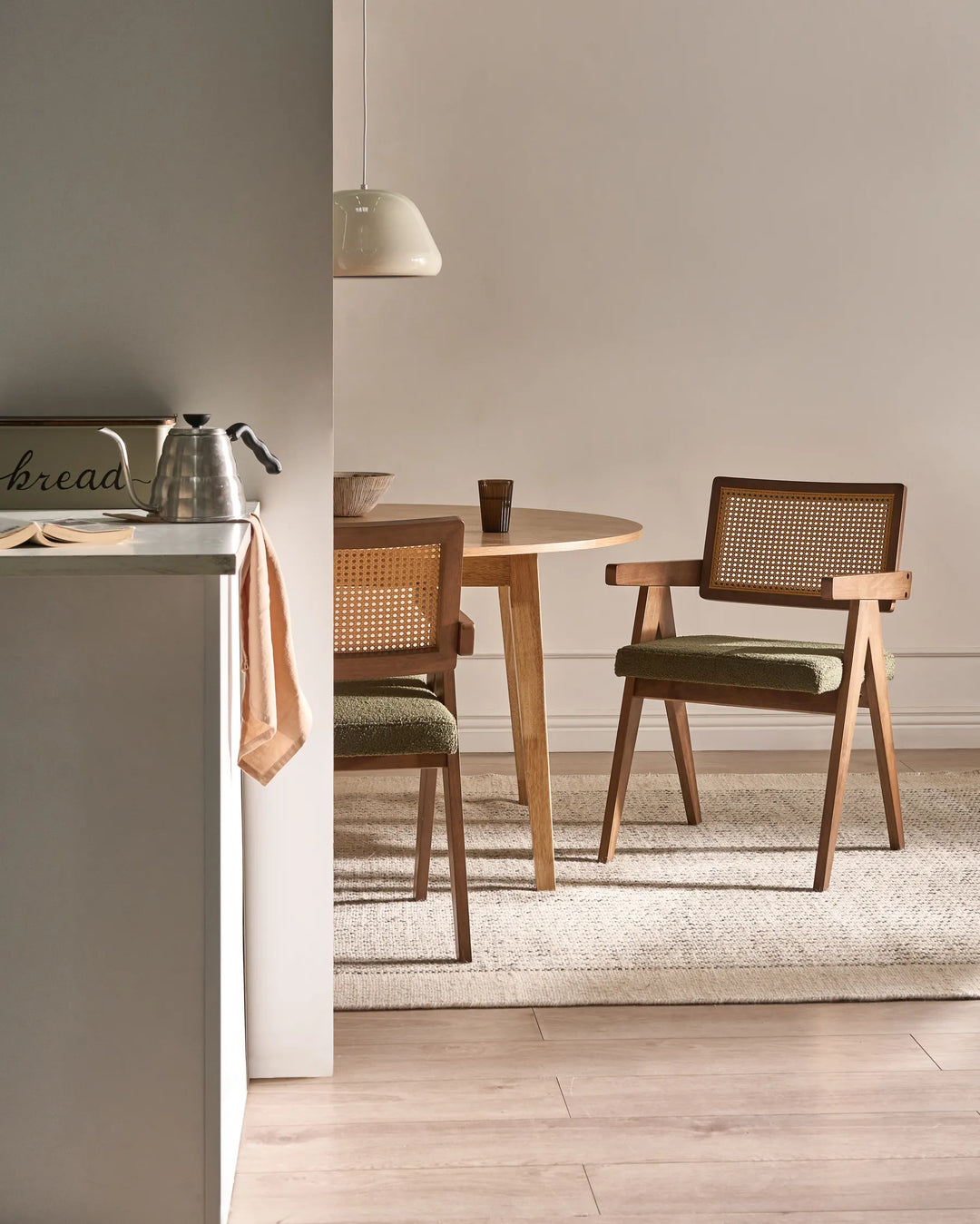 A mid-century modern wooden dining chair with a woven cane backrest and green cushioned seat, shown in a styled dining room setting next to a round wooden table and a light-colored rug.