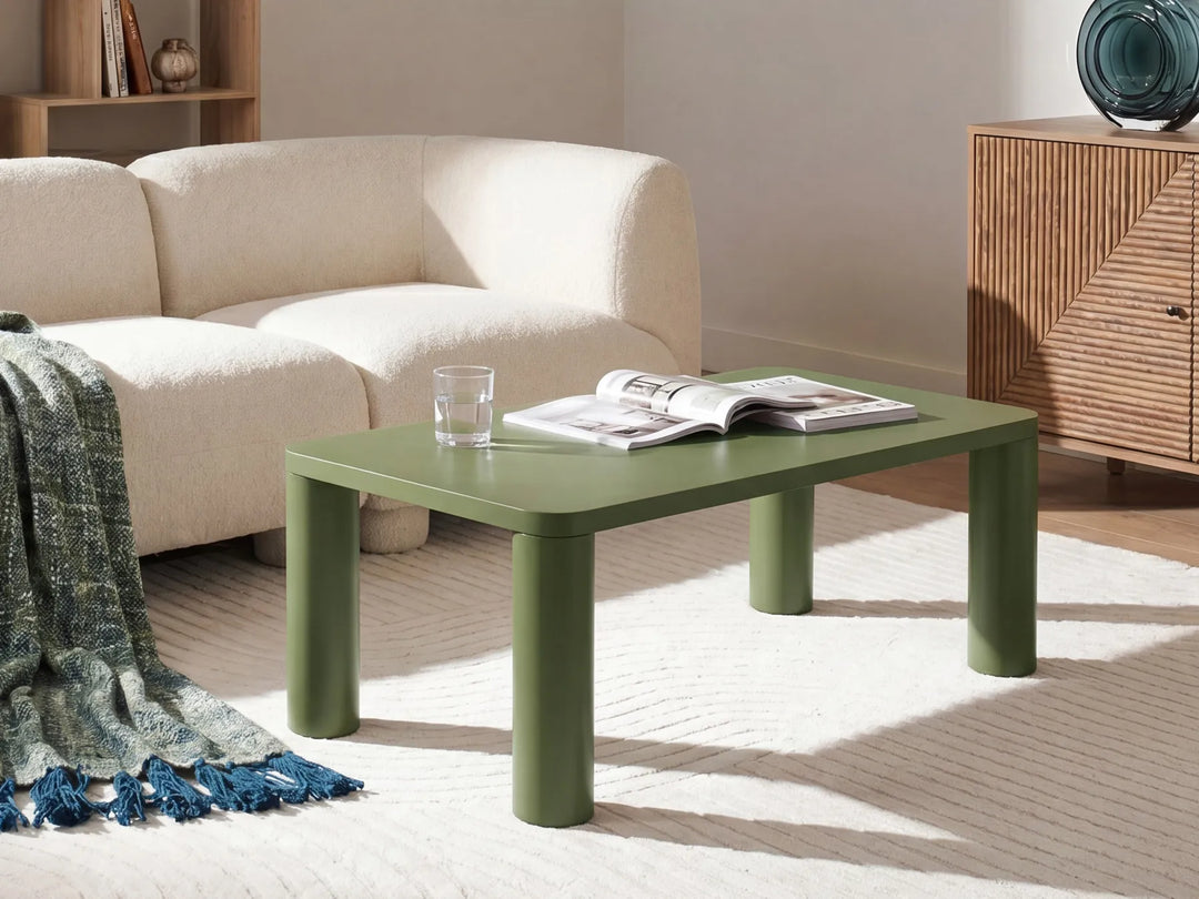 A low rectangular coffee table in sage green with thick cylindrical legs, styled in a modern living room setting with a cream sofa, a woven cabinet, and a light rug.