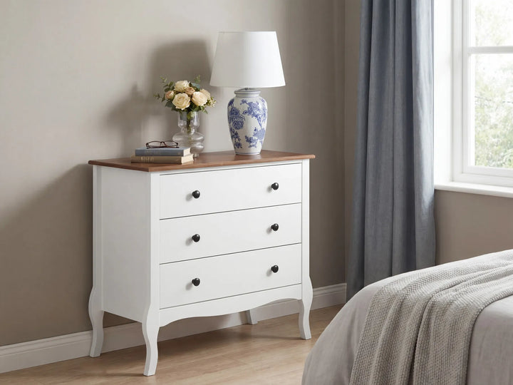 A white three-drawer nightstand with a natural wood top, styled in a bedroom setting next to a bed with gray curtains and a blue-and-white ceramic table lamp on top.