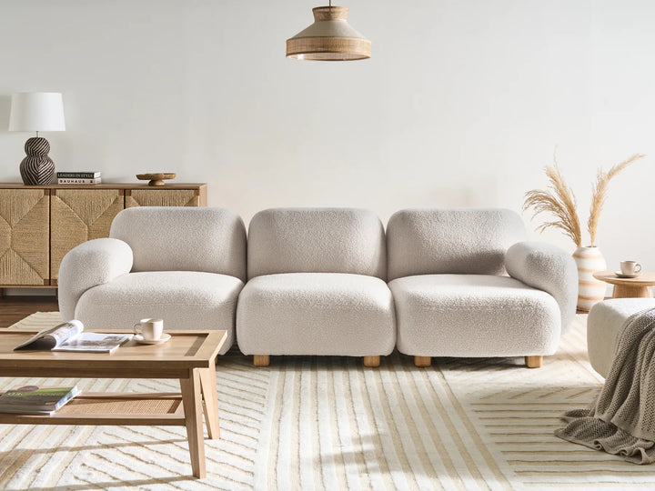 A modern three-seater sofa upholstered in a textured cream bouclé fabric, styled in a minimalist living room with a striped rug, wooden coffee table, and neutral decor.