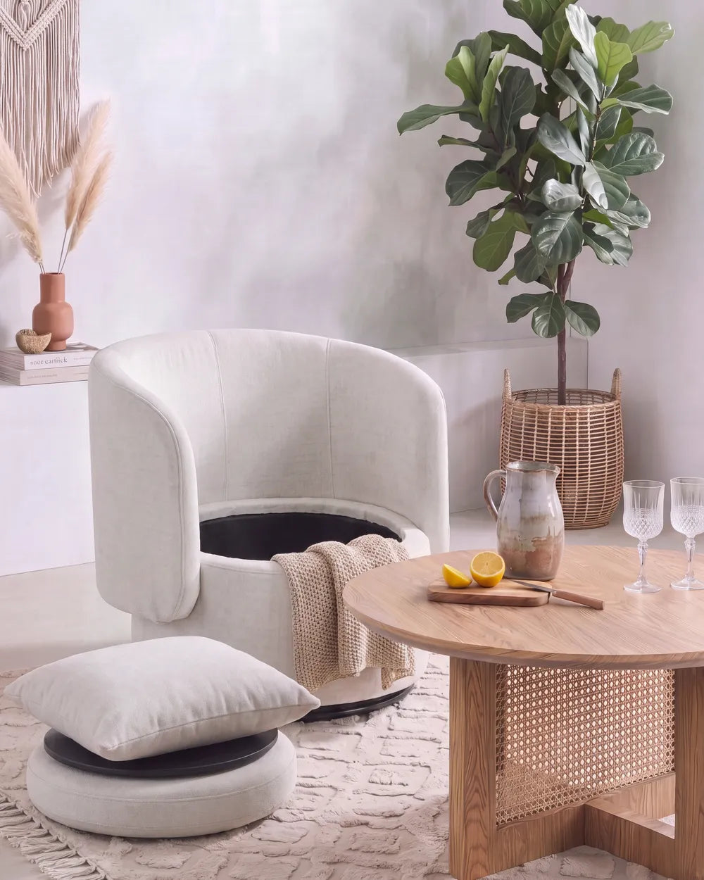 A cream upholstered barrel-style armchair with a matching cushion and throw blanket, styled in a bright, modern living room next to a round wooden coffee table.
