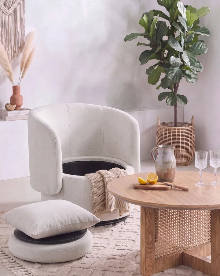 A cream upholstered barrel-style armchair with a matching cushion and throw blanket, styled in a bright, modern living room next to a round wooden coffee table.