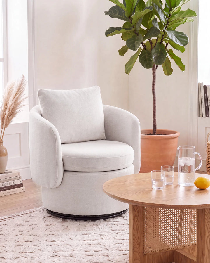 A light beige upholstered swivel armchair with a matching back cushion, placed in a bright, modern living room next to a wooden coffee table and a potted fiddle-leaf fig plant.