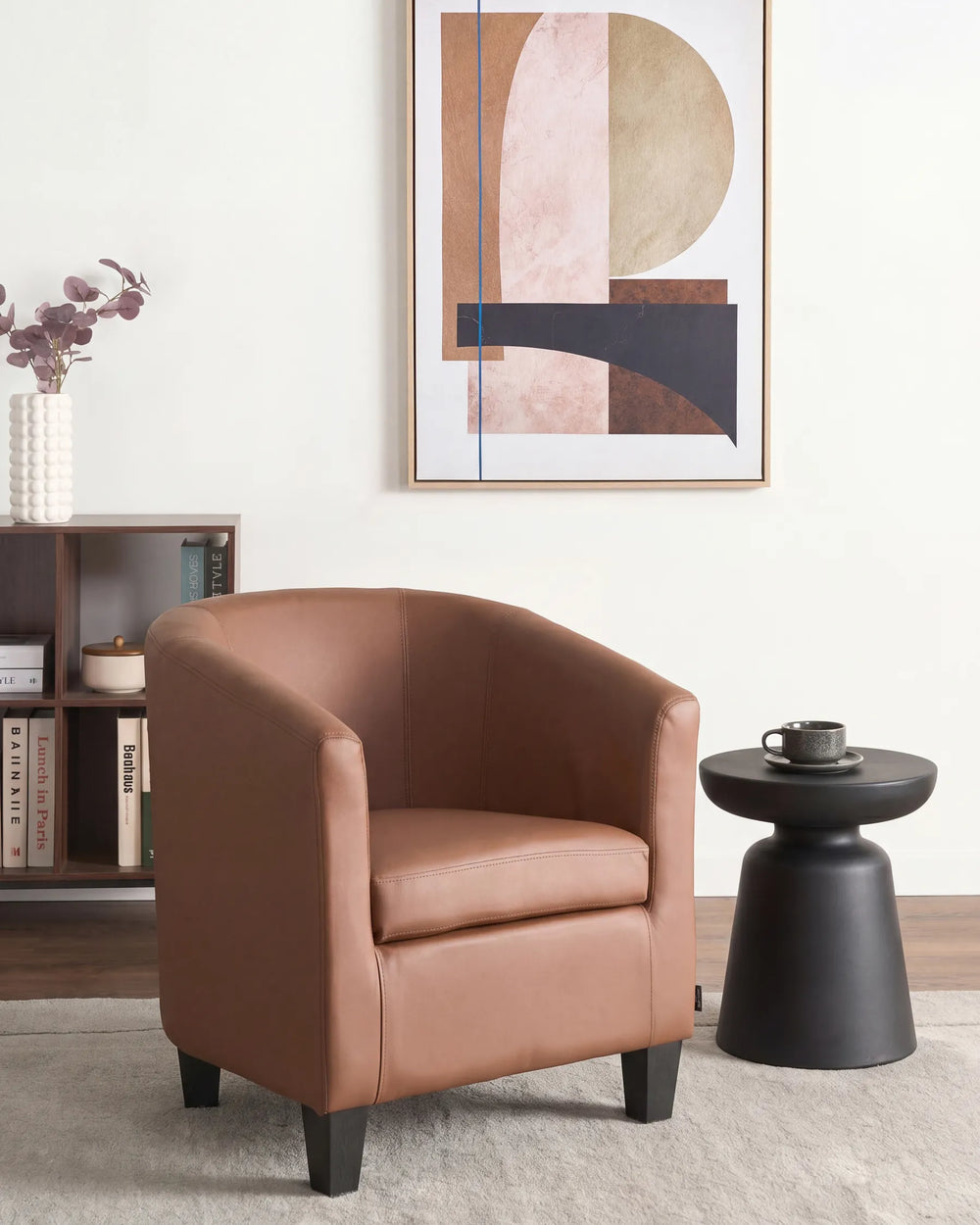 A modern barrel-style armchair upholstered in warm tan leather, styled in a minimalist living room next to a black side table and under abstract wall art.
