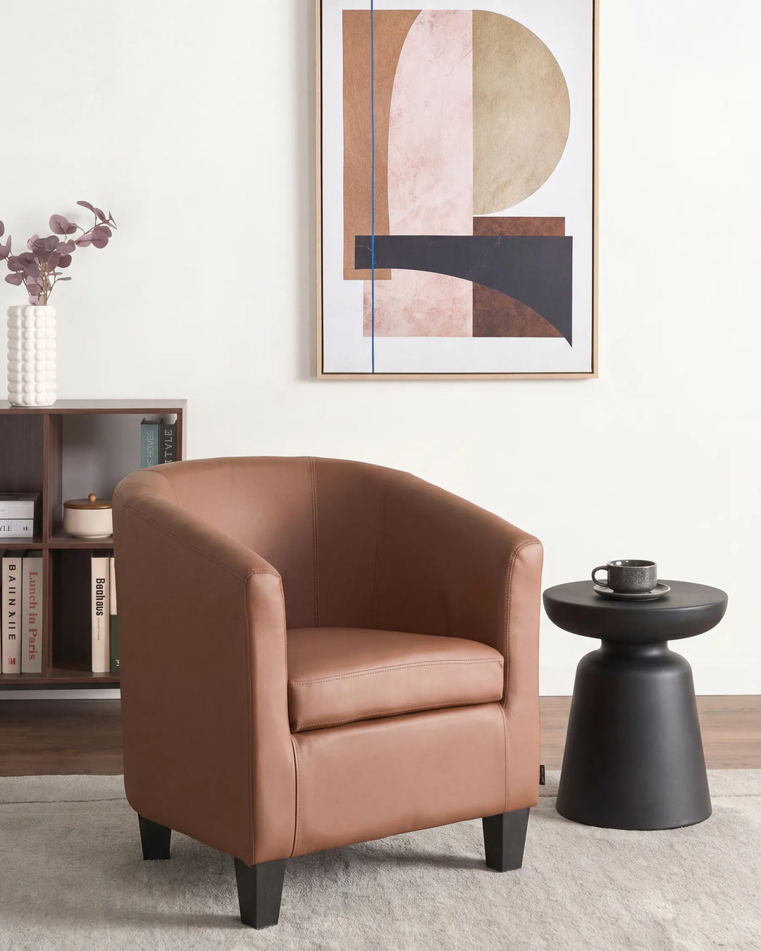 A modern barrel-style armchair upholstered in warm tan leather, styled in a minimalist living room next to a black side table and under abstract wall art.