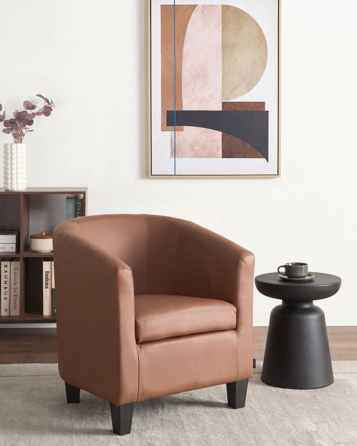 A modern barrel-style armchair upholstered in warm tan leather, styled in a minimalist living room next to a black side table and under abstract wall art.