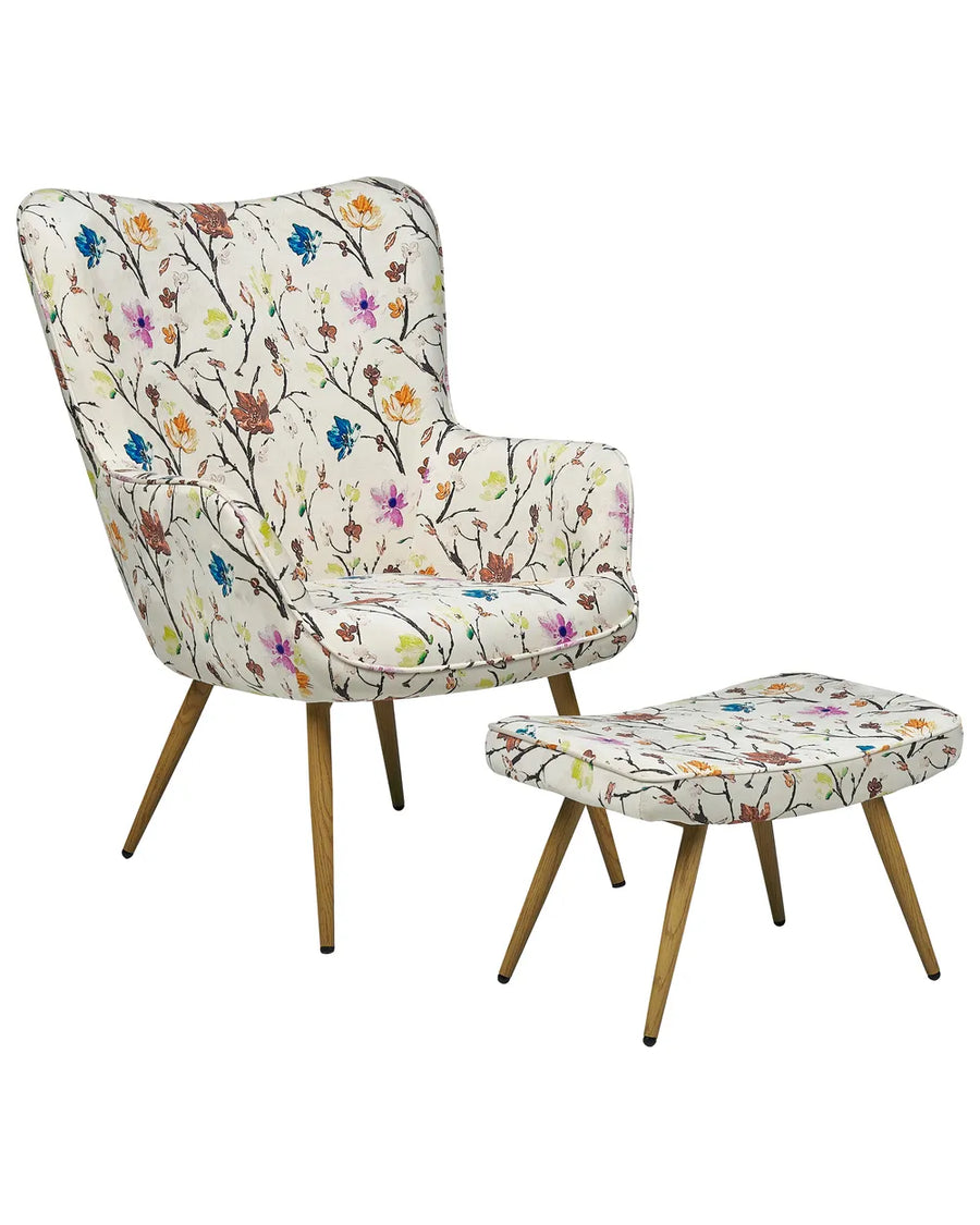 A mid-century modern wingback armchair with a matching ottoman, both upholstered in a cream fabric with a colorful floral and branch pattern, supported by angled wooden legs.